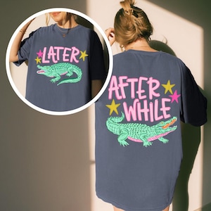 Peut inclure: T-shirt bleu marine avec un motif d'alligator vert et le texte "LATER" à l'avant et "AFTER WHILE" au dos. Le texte est rose et blanc avec des étoiles jaunes et roses. L'alligator a des accents roses.