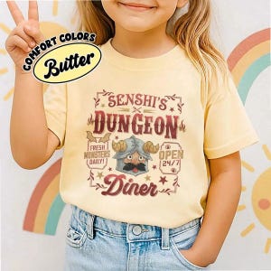 May include: A light yellow t-shirt with the text "SENSHI'S DUNGEON Diner" in a vintage style. The shirt features a cartoon character with a helmet and mustache. The words "Fresh Monsters Daily!" and "Open 24/7" are also printed on the shirt.