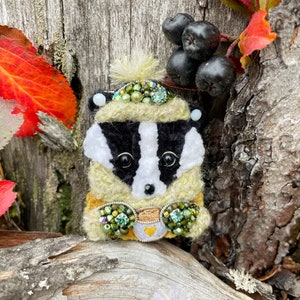 May include: A whimsical badger brooch with a yellow and green sweater, a cup of tea, and a white hat with green and black embellishments. The brooch is set against a backdrop of a wooden surface and autumn leaves.