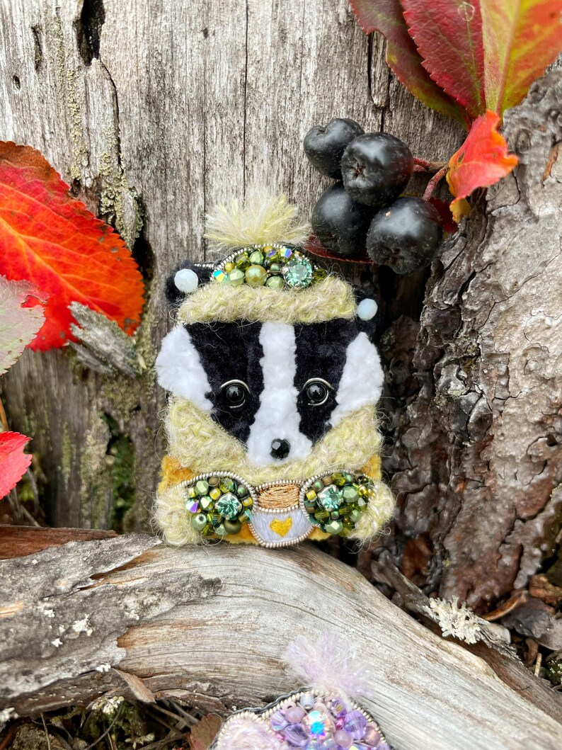 May include: A whimsical badger brooch with a yellow and green sweater, a cup of tea, and a white hat with green and black embellishments. The brooch is set against a backdrop of a wooden surface and autumn leaves.