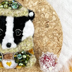 May include: A black and white badger wearing a green and gold embroidered hat and scarf. The badger is made of felt and has a beaded design.
