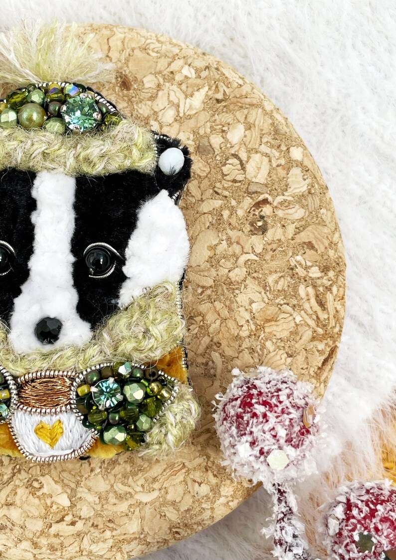 May include: A black and white badger wearing a green and gold embroidered hat and scarf. The badger is made of felt and has a beaded design.