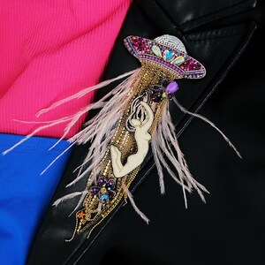 May include: A beaded brooch with a pink and purple UFO design, a white figure, and pink feathers.