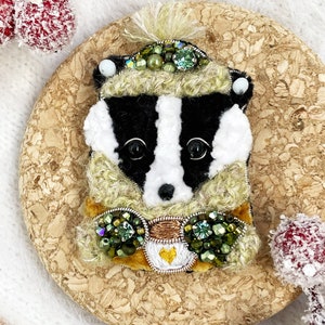 May include: A badger wearing a green and white hat and scarf with green and gold embellishments. The badger is holding a small cup with a heart on it.