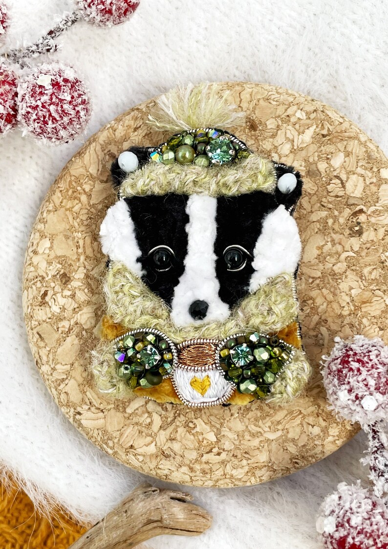 May include: A badger wearing a green and white hat and scarf with green and gold embellishments. The badger is holding a small cup with a heart on it.