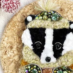 May include: A black and white badger wearing a green and gold embroidered hat and scarf. The badger is holding a small cup with a heart on it.