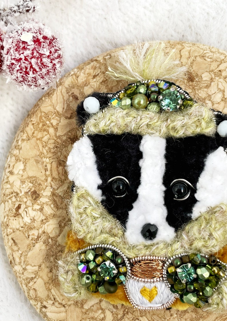 May include: A black and white badger wearing a green and gold embroidered hat and scarf. The badger is holding a small cup with a heart on it.