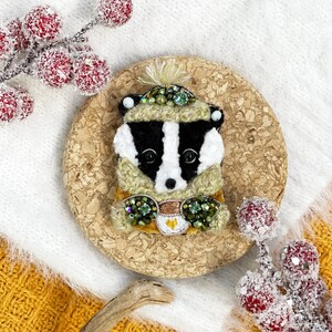 May include: A badger wearing a green and yellow hat and scarf, holding a cup of coffee. The badger is made of felt and decorated with beads and sequins. The badger is sitting on a cork coaster.