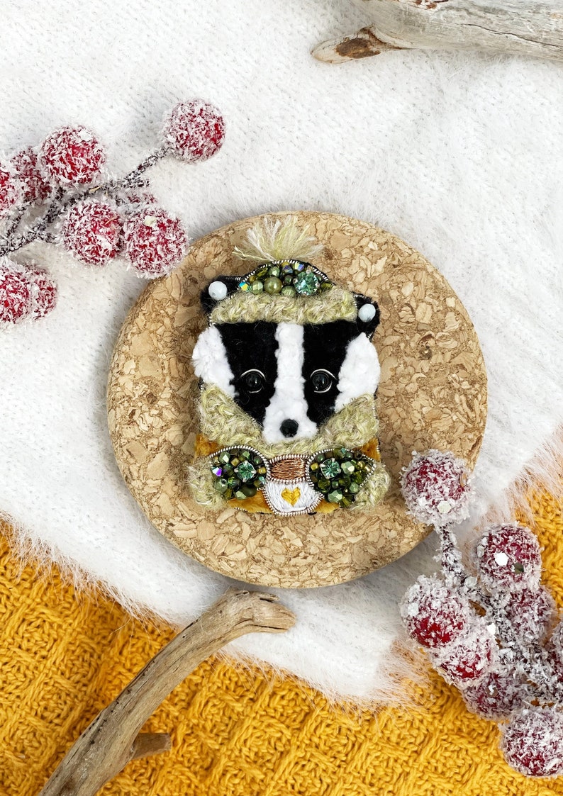 May include: A badger wearing a green and yellow hat and scarf, holding a cup of coffee. The badger is made of felt and decorated with beads and sequins. The badger is sitting on a cork coaster.