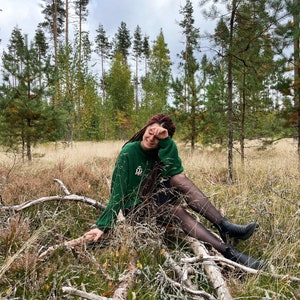 May include: A woman with red hair sits on a fallen log in a forest. She is wearing a green sweater, black tights, and black boots. The forest floor is covered in brown leaves and pine needles.