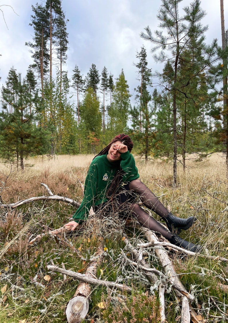 May include: A woman with red hair sits on a fallen log in a forest. She is wearing a green sweater, black tights, and black boots. The forest floor is covered in brown leaves and pine needles.