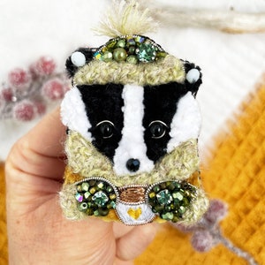 May include: A black and white badger wearing a green and brown crocheted hat with green and gold embellishments. The badger is holding a small white cup with a gold heart.