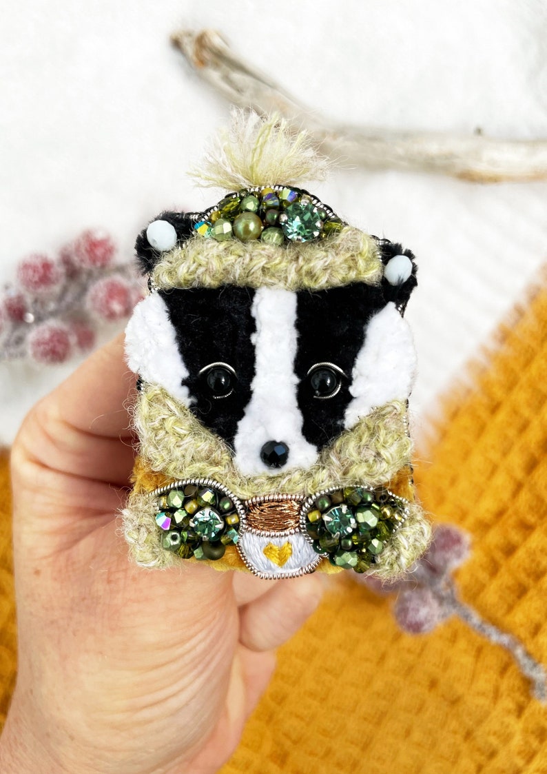 May include: A black and white badger wearing a green and brown crocheted hat with green and gold embellishments. The badger is holding a small white cup with a gold heart.