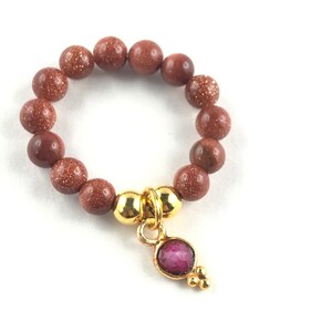 Könnte beinhalten: Ein Ring mit rötlich-braunen, gesprenkelten Perlen. Ein goldfarbener Anhänger mit einem kleinen, ovalen, rosa Edelstein und drei kleineren goldenen Perlen hängt an dem Ring. Der Ring ist vor einem weißen Hintergrund platziert.