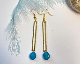 Könnte beinhalten: Ein Paar goldfarbene Ohrringe mit rechteckigem Design und einem blauen Stein am unteren Ende. Die Ohrringe liegen auf einer weißen Oberfläche mit einer hellblauen Feder und einer Muschel im Hintergrund.