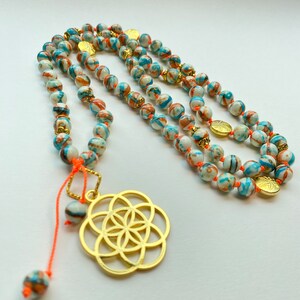 Könnte beinhalten: Eine weiße und blaue Perlenkette mit goldenen Akzenten und einem goldenen Lebensblume-Anhänger. Die Kette hat eine orange Schnur, die am Anhänger befestigt ist.