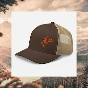 Può includere: Cappellino da camionista marrone con retro in rete beige. La parte anteriore del cappello presenta un design di corna di cervo ricamate arancioni. Il cappello ha una visiera curva e una chiusura a scatto.
