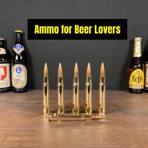 Puede incluir: Una disposición de botellas de cerveza y abridores de botellas con forma de bala. El texto "Ammo for Beer Lovers" se muestra encima de los artículos. Los abridores de botellas son de color dorado y están colocados delante de las botellas de cerveza.