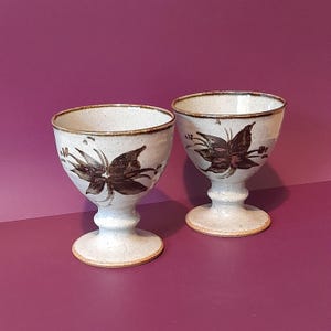 May include: A pair of off-white ceramic goblets featuring a brown floral pattern. Each goblet has a pedestal base and a wide rim. The floral design is painted on the front. The background is a solid purple colour.