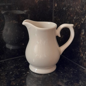 May include: A solid off-white ceramic cream jug featuring a rounded body, a curved handle, and a small pouring spout. The jug has a smooth, glossy finish and sits on a circular base, showcasing a simple and functional design.