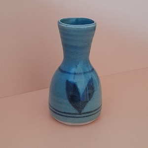 May include: A small, turquoise ceramic vase with a narrow neck and rounded body. The vase features a dark blue heart-shaped design and two horizontal lines. The vase is displayed against a light pink background.