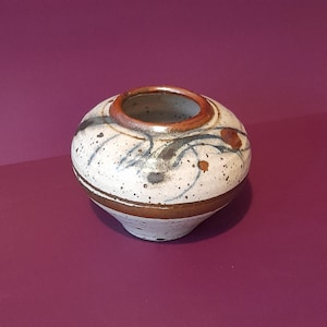 May include: A small, round ceramic vase with a speckled white body. The vase has brown bands at the base and rim, with decorative blue and brown brushstrokes. The interior is a darker brown colour.
