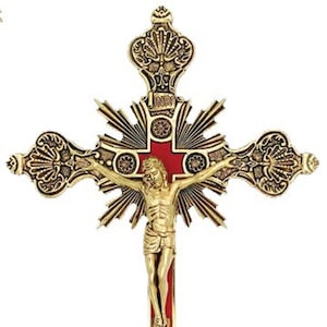 May include: A gold-toned metal crucifix with a red center and intricate details. The figure of Jesus Christ is depicted in a realistic style. The crucifix is mounted on a decorative stand with a three-pronged base.