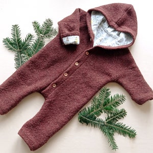 Könnte beinhalten: Burgunderfarbener Baby-Overall mit Kapuze und Holzknöpfen. Die Innenseite der Kapuze und der Manschetten hat einen hellblauen Stoff mit floralem Muster. Der Overall ist aus einem strukturierten Material. Tannenzweige sind um das Kleidungsstück angeordnet.