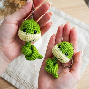 Op de afbeelding: Twee handgemaakte gehaakte sleutelhangers in de vorm van groen-crèmekleurige wezens. De sleutelhangers hebben zwarte ogen en worden in open handen gehouden. De achtergrond is wazig, met de focus op de sleutelhangers.