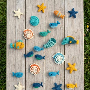 May include: A selection of handmade crochet sea creatures, including starfish, fish, whales, and seashells, in shades of blue, yellow, and orange. The items are arranged on a weathered wooden surface, creating a nautical theme.