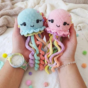 Puede incluir: Dos juguetes de medusas de ganchillo hechas a mano, uno azul claro y otro rosa, cada uno con caras sonrientes y tentáculos coloridos. Las medusas se sostienen en manos abiertas, mostrando su diseño caprichoso. Se ve un reloj con una correa pastel.