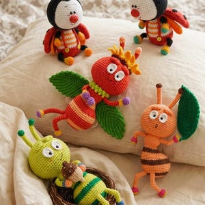 Puede incluir: Una colección de juguetes de insectos hechos a mano con ganchillo en varios colores. Los juguetes incluyen una mariquita, una abeja, una mosca con alas verdes y una oruga en un nido. Los juguetes están hechos de hilo y están sobre una almohada blanca.