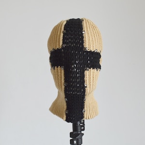 Könnte beinhalten: Beige gestrickte Sturmhaube mit einem großen schwarzen Kreuzmuster auf der Vorderseite. Die Sturmhaube hat kleine silberne Nieten und wird auf einem schwarzen Ständer präsentiert. Die Kopfbedeckung besteht aus einem weichen, strukturierten Material.