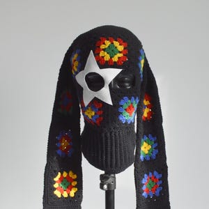 May include: Black crochet balaclava with a white star-shaped eye covering. The balaclava features colorful floral granny squares and long ear-like extensions. The design incorporates red, yellow, blue, and green accents.