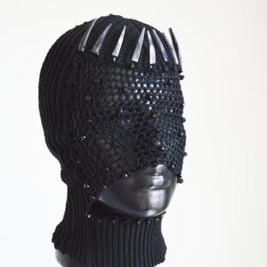 May include: A black crocheted balaclava featuring a dense ribbed knit base and an open-weave mesh face covering. The headpiece is adorned with a row of pointed, silver-toned metallic accents across the forehead and small black beads.