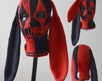 Könnte beinhalten: Eine schwarze und rote Häkel-Balaclava mit langen Hasenohren. Die Balaclava hat ein aufgenähtes Gesicht mit einem bedrohlichen Ausdruck.