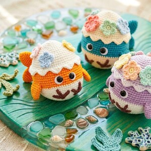 May include: Three handmade crochet fish toys in orange, blue, and purple, each adorned with floral accents. The fish have large googly eyes and are set on a decorative tray with sea glass and shells.