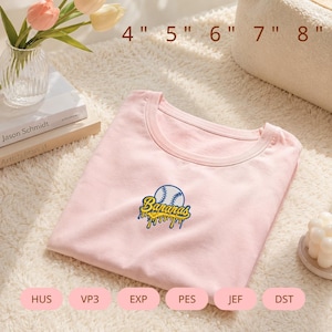 Puede incluir: Una camiseta rosa claro con un bordado central de una pelota de béisbol y la palabra Bananas en letra amarilla con un efecto de goteo azul. La imagen incluye opciones de tamaño de 4, 5, 6, 7, 8 pulgadas y formatos HUS, VP3, EXP, PES, JEF, DST.