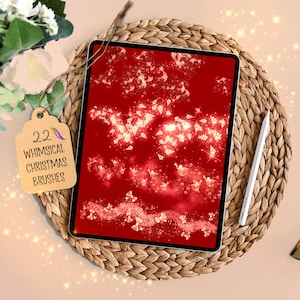 May include: A digital tablet shows a red background with white Christmas-themed graphics. A tag reads "22 Whimsical Christmas Brushes." A white stylus lies beside the tablet on a woven mat. The image has a festive, holiday theme.