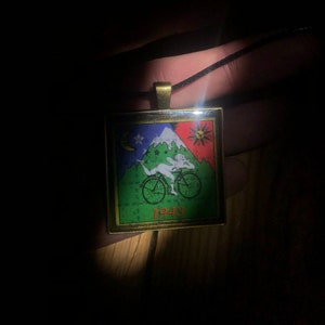 Peut inclure: Un collier avec un pendentif doré en forme de carré, orné d'une image colorée d'une personne à vélo avec une montagne et des étoiles en arrière-plan. Le pendentif porte également le texte "1943".