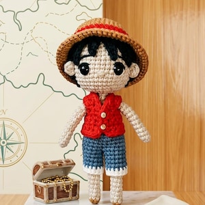 Pode incluir: Boneca de crochê de um personagem usando um chapéu de palha, colete vermelho e shorts azuis. A boneca está em uma superfície de mármore com um baú do tesouro e uma âncora por perto. Um mapa está ao fundo.