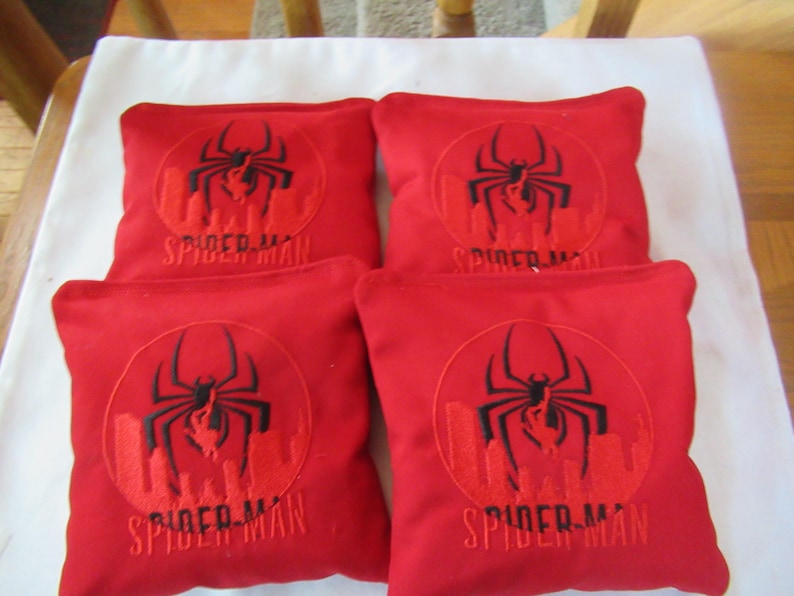 May include: Four red bean bag toss game bags. Each bag features a black spider design inside a circle, with the words "SPIDER-MAN" below. The bags are square and appear to be made of a soft fabric.