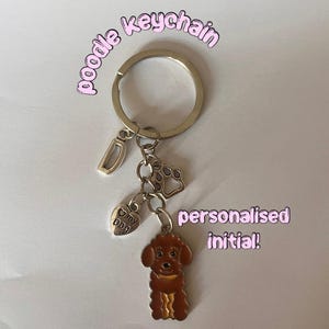 May include: A silver keychain featuring a brown poodle charm, paw print charm, heart charm, and a letter "D" charm. The poodle has a yellow collar. Text on the image says "poodle keychain" and "personalised initial!"