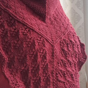 Könnte beinhalten: Ein weinroter Strickschal mit einem strukturierten Muster. Der Schal ist über den Schultern drapiert und zeigt das komplizierte Design und die satte Farbe. Die Kanten haben einen gewellten Abschluss.