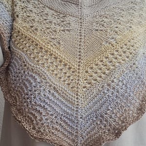 Könnte beinhalten: Ein dreieckiges, handgestricktes Tuch mit einem komplizierten Spitzenmuster. Das Kleidungsstück zeigt einen Farbverlauf, der von einem blassen Gelb am Halsausschnitt zu einem sanften Lavendel übergeht und an der Bogenkante in Braun endet.