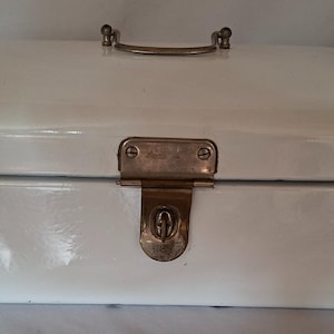 May include: A rectangular off-white enameled metal bread box with rounded corners. The container features a brass-toned metal handle on the top lid and a central brass-toned latch mechanism on the front for securing the lid in a closed position.