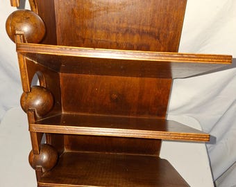 Peut inclure: Une étagère murale en bois composée de trois niveaux horizontaux. Le côté gauche est soutenu par une structure verticale intégrant trois éléments décoratifs sphériques en bois. Deux crochets de suspension en métal sont fixés au sommet.