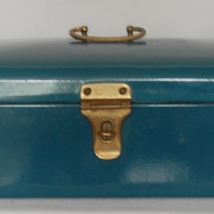 May include: Teal-colored rectangular box with a rounded top. The box features a brass-colored handle and a clasp closure. The box is likely used for storage or display of small items.