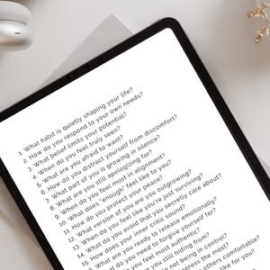 May include: A black tablet displays a list of self-reflection questions in white text. The tablet is on a white surface, with a pair of white headphones and a decorative plant in the background. The questions are about self-awareness and personal growth.