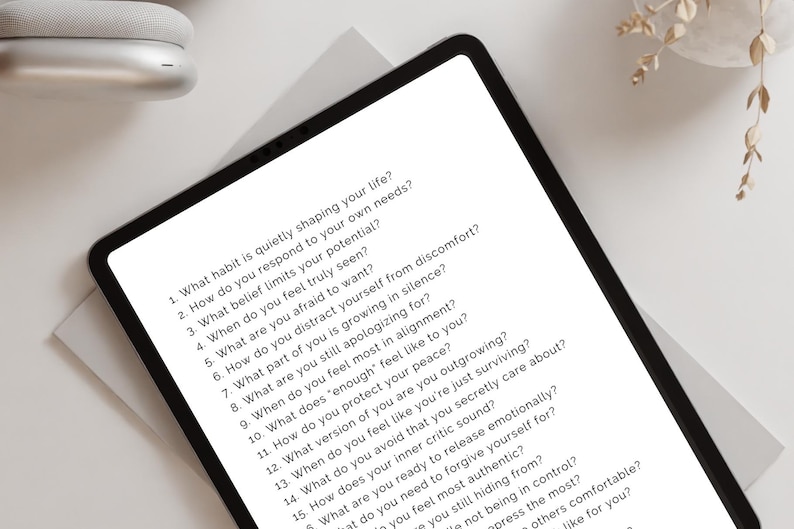 May include: A black tablet displays a list of self-reflection questions in white text. The tablet is on a white surface, with a pair of white headphones and a decorative plant in the background. The questions are about self-awareness and personal growth.